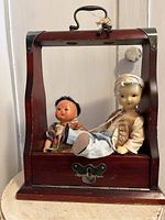 Full view of the two Asian character dolls seated inside the wood tantalus with handle and keyhole plate with keys.