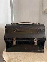 Front view of a black metal lunch box with a curved lid and two metal clasps, placed on a round table showcasing some surface wear and scratches.