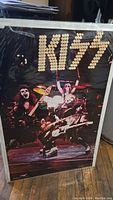 Shows full front of the Kiss band circa 1975 poster inside plastic sleeve, with the band performing on stage, wearing signature makeup and costumes.