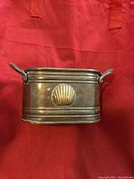 Side view of brass container showing shell motif and dual handles.