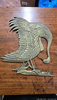 Full view of flat brass bird decorative piece on wooden table, showing detailed feathers and curved neck.