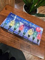 Box of Holiday Light Bulbs on wooden table with houseplant in background, showing colorful bulb images and product details on packaging.