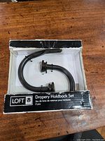 Photo of two black metal curved drapery holdbacks in original box on wooden surface, showing product and packaging.