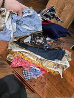 Hand holding stack of various vintage textile scraps showing different colors and prints such as gingham, denim, paisley, and floral patterns on a wooden table surface.