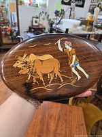 Oval wooden plaque held in hand showing entire inlaid art with two oxen and man.