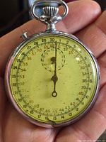 Front view of pocket stopwatch showing yellowed dial with numeric scales and single central hand.