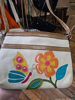 Front view of the off-white fabric purse with brown trim, decorated with a colorful butterfly and flower applique with studs and stitching.