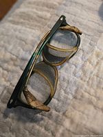 Side and top angled view of a single pair of vintage horn rimmed glasses with metal mesh side shields and flex fit temples lying on a soft surface.