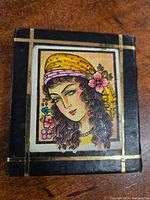 Front view of vintage hand mirror showing colorful hand-painted girl portrait with yellow headscarf, flowers, and gold trim on black leather background.