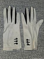 Pair of white leather gloves with black buttons on fingers, laid flat on gray surface.