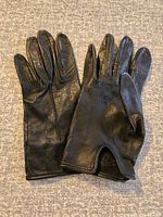 Pair of black leather gloves laid flat on a gray fabric surface. Gloves have a small notch near the wrist and slight sheen indicating leather material.