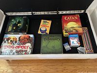 Photo shows seven assorted board games and puzzles including Catan, Dog-Opoly, Villainous, Brain Twisters book, Ravensburger puzzle, Kalvinet card game, and a wooden cribbage board with pegs and cards stored in a fabric bin.