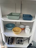 Shelf view showing two rectangular baking pans (green and blue) on top shelf, and six mixing bowls (blue, yellow, orange, four white) on middle shelf with toaster and spice rack below