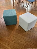 Two soft-sided cube stools, one blue-green and one cream, positioned on a wooden floor beside a dining table.
