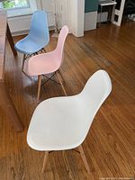 Three modern dining chairs, one white, one pink, and one blue, showing the side profile with wooden legs on hardwood floor.