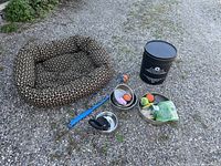 Photo shows dog bed, two metal bowls with toys, grooming brush, a black container, and dog treat packet on ground.