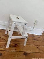 White two-step foot stool placed on a wooden floor, showing side view with visible bolts and a cutout handle on top.