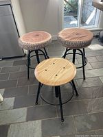 Three wood and metal kitchen stools, two with tree-ring and bark style cushions, one with plain wood seat, shown on tiled floor near window and fridge.