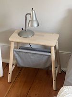 Wooden nightstand with grey fabric sling storage and silver metal table lamp on top, standing on wooden floor.