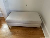 Upholstered ottoman/bench closed, showing light beige fabric and metal legs on wood floor.