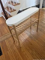 Side view of the oval side table showing wood frame, white plastic top and slatted lower shelf, placed on hardwood floor near staircase.