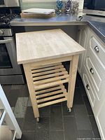 Side view of the Ikea wooden rolling cutting board stand placed on a tiled kitchen floor next to cabinetry and appliances, showing hardwood top and slatted shelves with caster wheels.