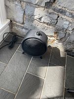 Black NOMA small ceramic space heater placed on tiled floor near stone wall, showing front grill and power cord.
