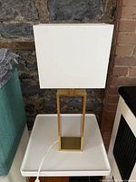 Front view of gold coloured table lamp with white rectangular shade placed on white tray against stone and brick wall background.