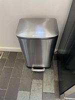 Front view of rectangular stainless steel trash bin with foot pedal on tiled floor near wall.