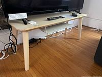 TV table with under shelf and TV on top, showing light wood surface, rounded corners, and white undershelf. Table on wooden floor with visible cords below.