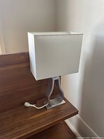 Side view of one lamp showing white rectangular fabric shade and curved silver metal base placed on a wooden surface.