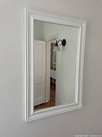 Photo of a rectangular wall mirror with a white border/frame mounted on a wall, showing clear reflection of room elements.