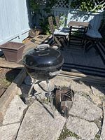 Weber kettle grill on tripod stand with two wheels outdoors on stone patio, charcoal chimney starter placed beside it.