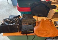 Six bags shown on a table including black reptile embossed vinyl purse with chain strap, brown leather bag, studded black purse, and other fabric bags.