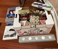 Partially open box on floor containing various candles and candle holders. Outer packaging includes Apollo branded box of long red tapered candles, several packs of tea lights, and decorative candle sets.