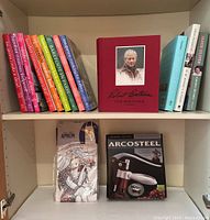 Shelving unit with 9 Ina Garten Barefoot Contessa cookbooks with colorful spines, positioned next to other cookbooks and Robert Bateman art book. Below are new apron in package and new Arcosteel corkscrew in box.