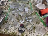 Multiple glass and crystal candleholders and coasters arranged on a floral cloth surface, showing variety of shapes and designs.
