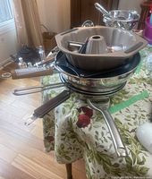 Stack of frying pans, Farberware bundt cake pan on top, stainless steel wok below them on floral tablecloth