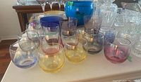 Front view of a group of 12 assorted colorful short drink glasses on a white tabletop with other glassware and dishes in the background.