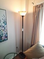 Full height view of the floor lamp standing in corner beside window and painting, lamp lit showing glass shade design.