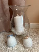 Photo showing the two Lladro porcelain bells with different ribbons and the glass hurricane candle holder with a white pillar candle inside on a carpeted floor.
