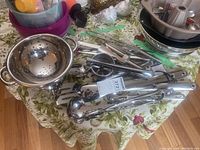Overview pile of mixed stainless steel kitchen utensils on floral tablecloth including ladles, slotted spoons, tongs, wire whisk, handheld strainer and other tools.