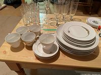 Photo showing set of cups with saucers, glasses, and stacked plates with gold floral motif on a wooden table.