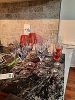 Photo showing a variety of stemware including clear wine glasses with black leaf design, red champagne flutes, and clear champagne flutes grouped together on a counter.