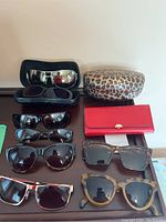 Photo showing 9 pairs of sunglasses displayed on a table with 3 sunglasses cases including a leopard print Guess case, a red case and a black case.