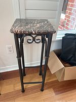 Front perspective view of the marble top table stand showing the marble surface and metal legs against a wooden floor and white wall.