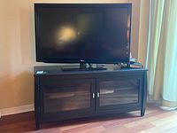 Front view of the black MDF TV stand cabinet with a TV on top, showing the two glass doors and silver handles.
