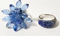 Two vintage costume rings displayed side by side on a white background, one large starburst style with blue marquise stones and one smaller silver-toned ring with blue rhinestones