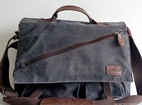 Full front view of the black resistant canvas RAVUO messenger bag showing diagonal zip pocket and carrying handle on flap.