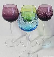 Front view showing four colored crystal goblets in purple, dark purple, blue, and green with cut patterns on stems and bowls